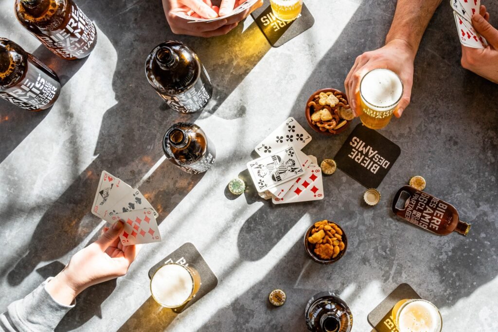 Shooting d'ambiance avec des personnes réunies autour d'une table, jouant aux cartes avec leur verre de Bière des amis Blonde (5,8% Alc.) accompagné de bouteilles de 33cl et 66cl de Bières des amis Blonde (5,8%).